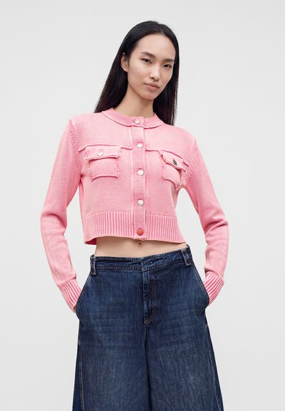 Jeune femme portant un gilet court rose boutonné avec des poches et un jean large bleu foncé, debout les mains dans les poches.