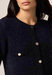 Cardigan navy in tessuto testurizzato con scollo rotondo, bottoni in hardware dorato e una tasca a toppa. Il tessuto ha un aspetto morbido e peloso.
