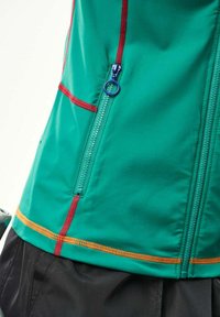 Zelená bunda s modrým ťahákom na zips, červeným šitím a oranžovým šitím na leme, nosená cez čierne nohavice.