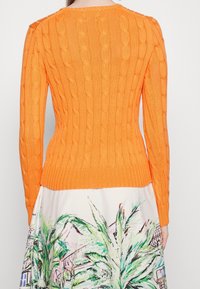 Nainen, jolla on päällään kirkkaan oranssi palmikkoneule ja valkoinen hame, jossa on värikäs abstrakti lehti- ja rakennuskuvio, näkyy takaapäin.