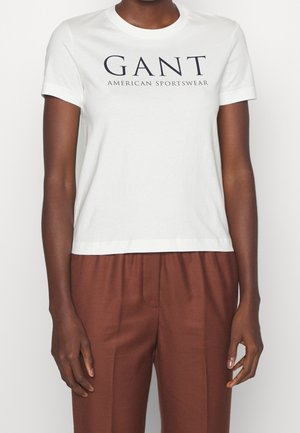 Person wearing a white GANT American Sportswear t-shirt and brown trousers, standing with arms relaxed by their sides.