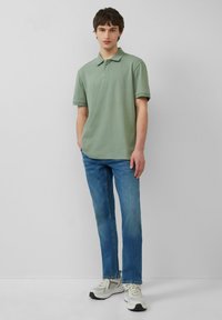 Camisa polo de algodón verde con cuello, tapeta de tres botones y mangas cortas, combinada con jeans de mezclilla azul y zapatillas ligeras.