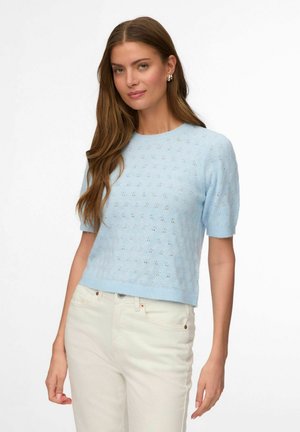 Jeune femme aux longs cheveux bruns portant un haut en maille bleu clair à manches courtes et un pantalon taille haute blanc cassé, debout devant un fond blanc.