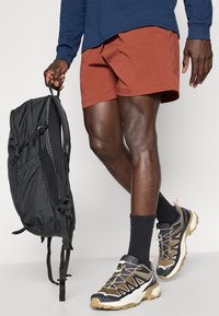 Schwarzer Rucksack, der mit einer Hand gehalten wird, kombiniert mit orangen Shorts, einem blauen Langarmshirt und robusten, mehrfarbigen Sportschuhen mit weißen, schwarzen und beigen Akzenten.