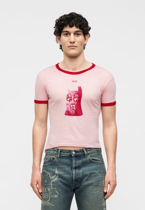 Mladý muž s kučeravými vlasmi nosí ružové krátke tričko s červeným lemom a grafickou potlačou tváre, v kombinácii s modrými džínsami s umeleckým motívom lebky.