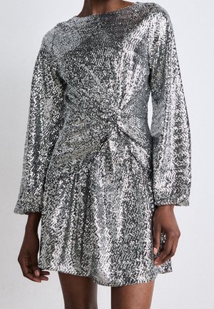 Mini-robe à sequins argentés scintillants avec manches longues et détail drapé subtil à la taille, portée par une personne sur un fond uni.