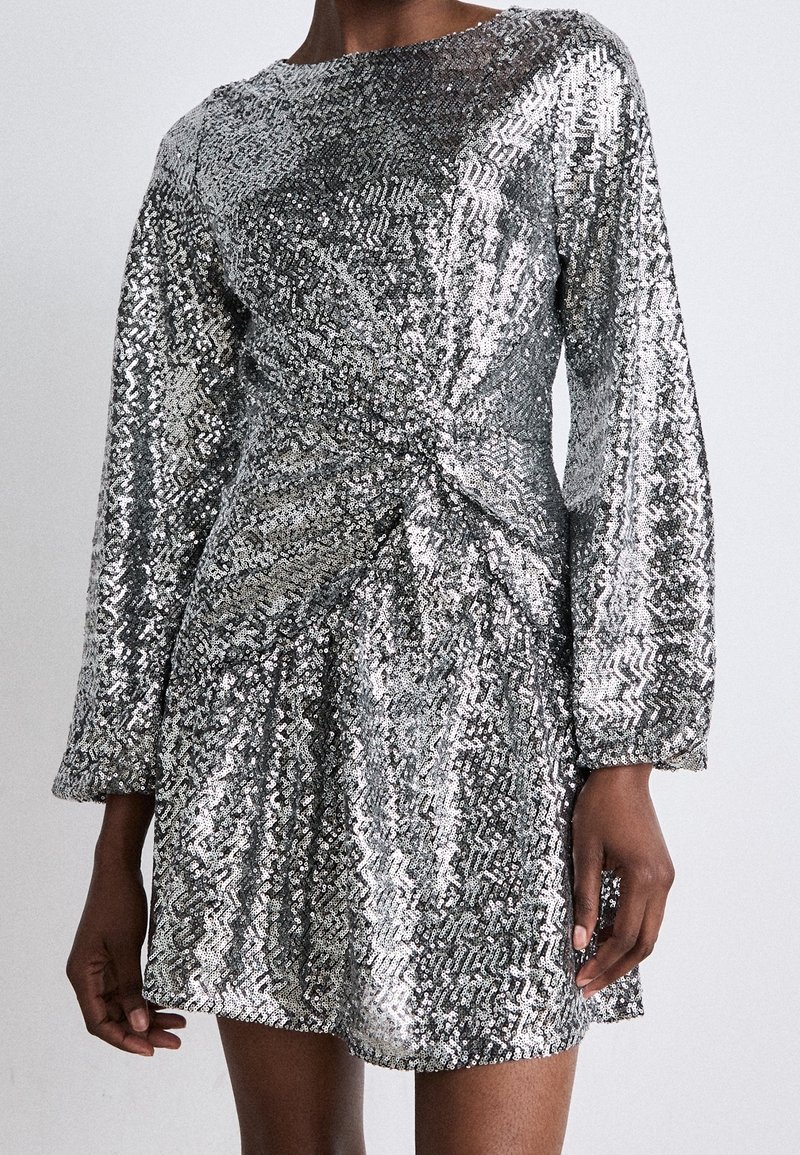 Mini-robe à sequins argentés scintillants avec manches longues et détail drapé subtil à la taille, portée par une personne sur un fond uni.