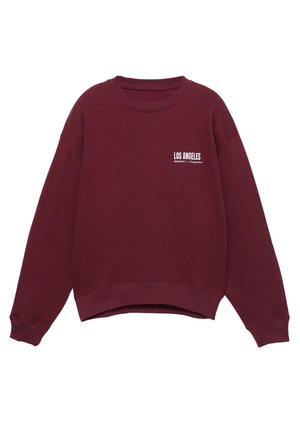 Personne portant un sweat-shirt bordeaux avec le texte "Los Angeles" sur une chemise aux rayures bleues et blanches et un pantalon noir, visible du cou jusqu’aux hanches.