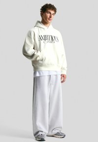 Fiatal férfi, aki túlméretezett fehér kapucnis pulóvert visel "AMBIOUS Fouts" felirattal, széles szürke melegítőnadrágban és szürke sportcipőben, sima háttér előtt áll.