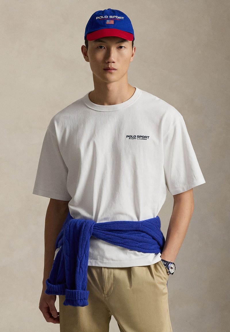 Λευκό βαμβακερό T-shirt με λογότυπο "POLO SPORT", σε συνδυασμό με μπλε πλεκτό πουλόβερ δεμένο γύρω από τη μέση, μπεζ παντελόνι, μπλε και κόκκινο καπέλο.