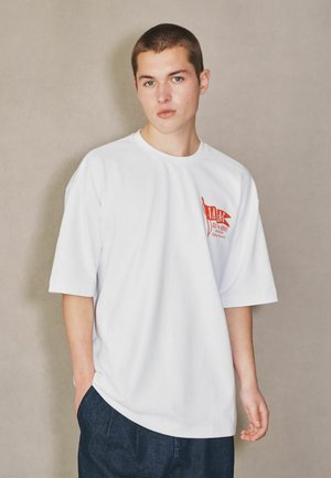 Persoană tânără cu păr scurt purtând un tricou alb larg cu un logo roșu pe piept și pantaloni bleumarin, stând pe un fundal simplu.