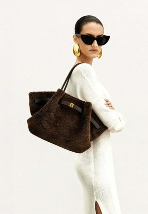 Femme en robe blanche en maille, portant de grandes boucles d'oreilles dorées et des lunettes de soleil noires style œil de chat, avec un grand sac fourre-tout marron pelucheux sur l'épaule.