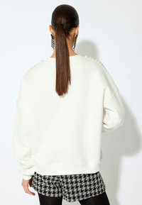 Sudadera blanca de gran tamaño con cuello redondo, tela texturizada y acentos de perlas a lo largo de los hombros. Llevada sobre pantalones cortos negros con estampado.