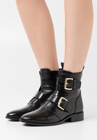 Botas de tobillo de cuero negro con una textura de cocodrilo brillante, que presentan dos hebillas doradas y un tacón bajo y cuadrado.