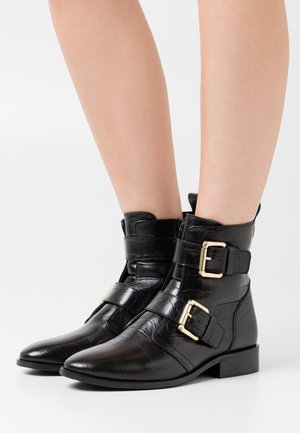 Botas de tobillo de cuero negro con una textura de cocodrilo brillante, que presentan dos hebillas doradas y un tacón bajo y cuadrado.