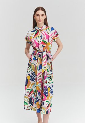Woman standing with hands in pockets wearing a colorful, short-sleeve, buttoned midi dress with a tie belt and abstract floral pattern.
