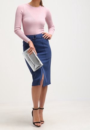 Donna che indossa una maglia rosa a maniche lunghe e una gonna a matita blu con cintura e spacco frontale, che tiene in mano una pochette argento e indossa tacchi neri con cinturino alla caviglia.