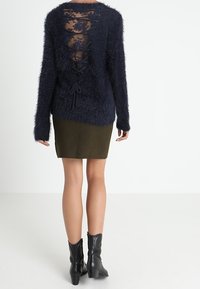 Marineblauer flauschiger Pullover mit transparentem Spitzeneinsatz am Rücken und Schleifendetails. Kombiniert mit einem grünen Rock und schwarzen Ankle-Boots.