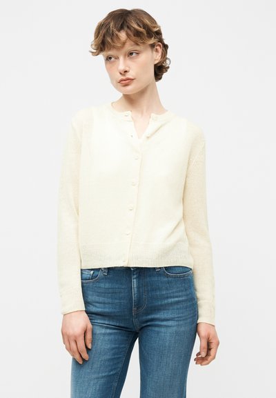 Νεαρή γυναίκα που φοράει μια κρεμ ζακέτα με κουμπιά και μπλε τζιν, στέκεται μπροστά σε λευκό φόντο, κοιτάζοντας προς το πλάι.