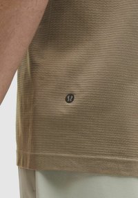 Beige Kurzarm-T-Shirt für den Sport mit strukturiertem Stoff, ausgestattet mit einem kleinen schwarzen Logo auf der unteren linken Seite.