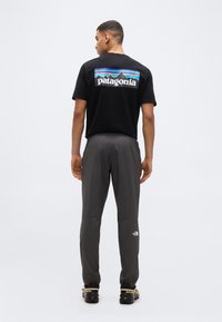 Άνδρας στέκεται με την πλάτη, φορώντας μαύρο t-shirt Patagonia, γκρι παντελόνι The North Face και μαύρα αθλητικά παπούτσια Salomon, σε λευκό φόντο.