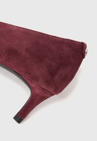 Chaussure à talon haut en daim bordeaux avec un bout pointu, un talon fin et une semelle noire ; présente des coutures discrètes et un accent métallique.