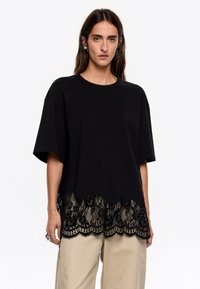 Must astumine lühikeste varrukatega must T-särk, mille alumises ääres on laineline pits. Pitsil on lillemuster, mis lisab materjalile tekstuuri ja kontrasti.