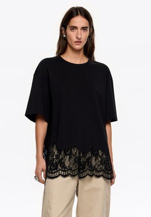 Camiseta negra de manga corta, con un borde de encaje en forma de scallop. El encaje tiene un estampado floral, añadiendo textura y contraste a la tela.