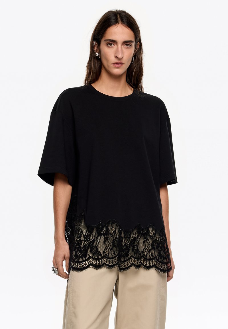 Must astumine lühikeste varrukatega must T-särk, mille alumises ääres on laineline pits. Pitsil on lillemuster, mis lisab materjalile tekstuuri ja kontrasti.