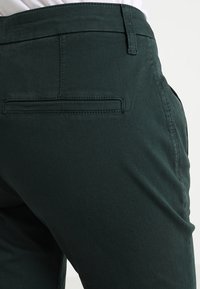 Pantaloni in cotone verde scuro con una tasca posteriore e dettagli di cucitura discreti. Texture liscia, design aderente con un girovita a media altezza.