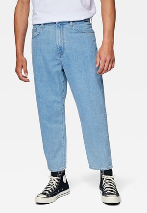 Personne portant un jean droit bleu clair, des baskets montantes noires avec des lacets blancs et une chemise blanche, debout sur un fond blanc.