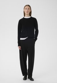 Črni pleteni pulover čez belo majico, združeni z ohlapnimi črnimi hlačami in črnimi čevlji, na nevtralnem ozadju.