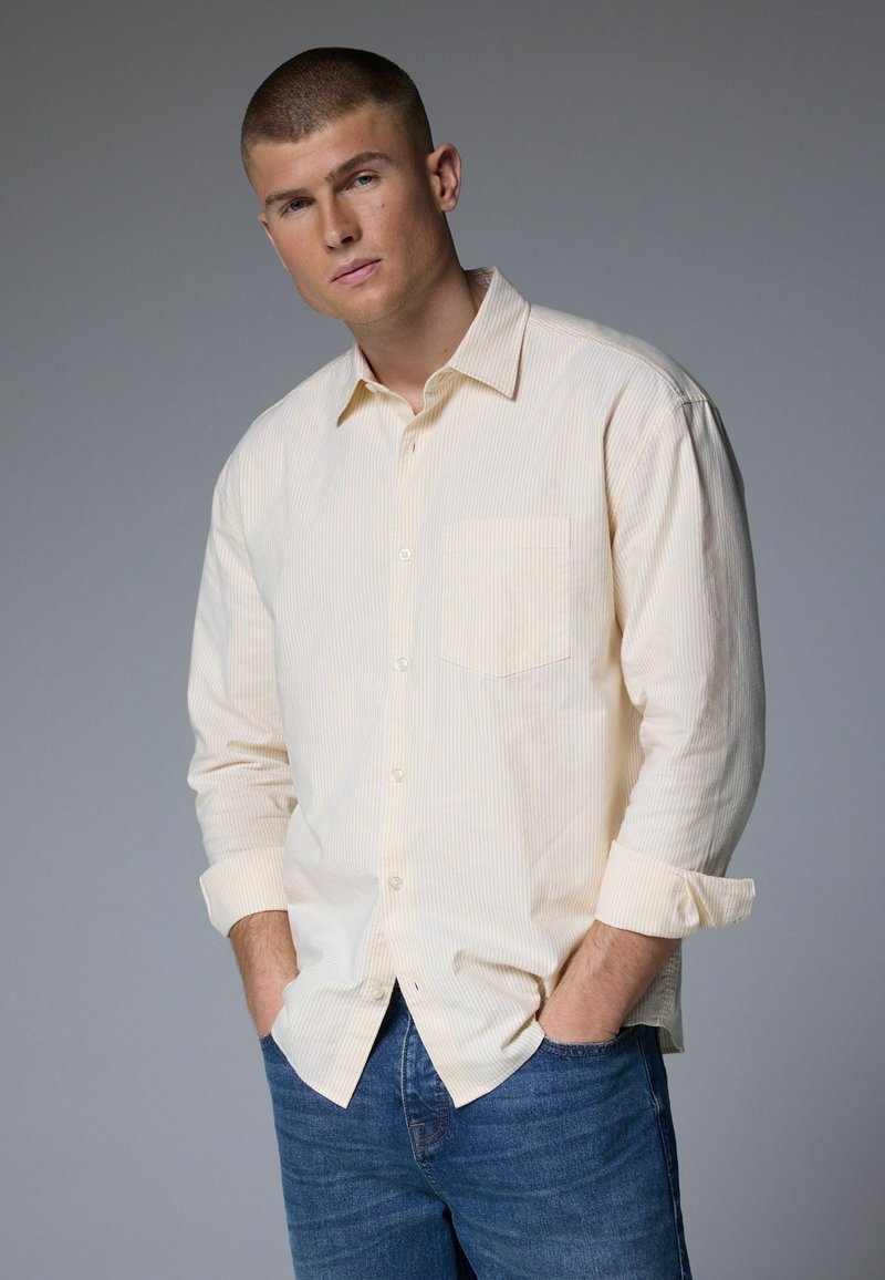 Light yellow striped shirt with a button-down collar, single chest pocket, and long sleeves rolled up. Worn with blue denim jeans.