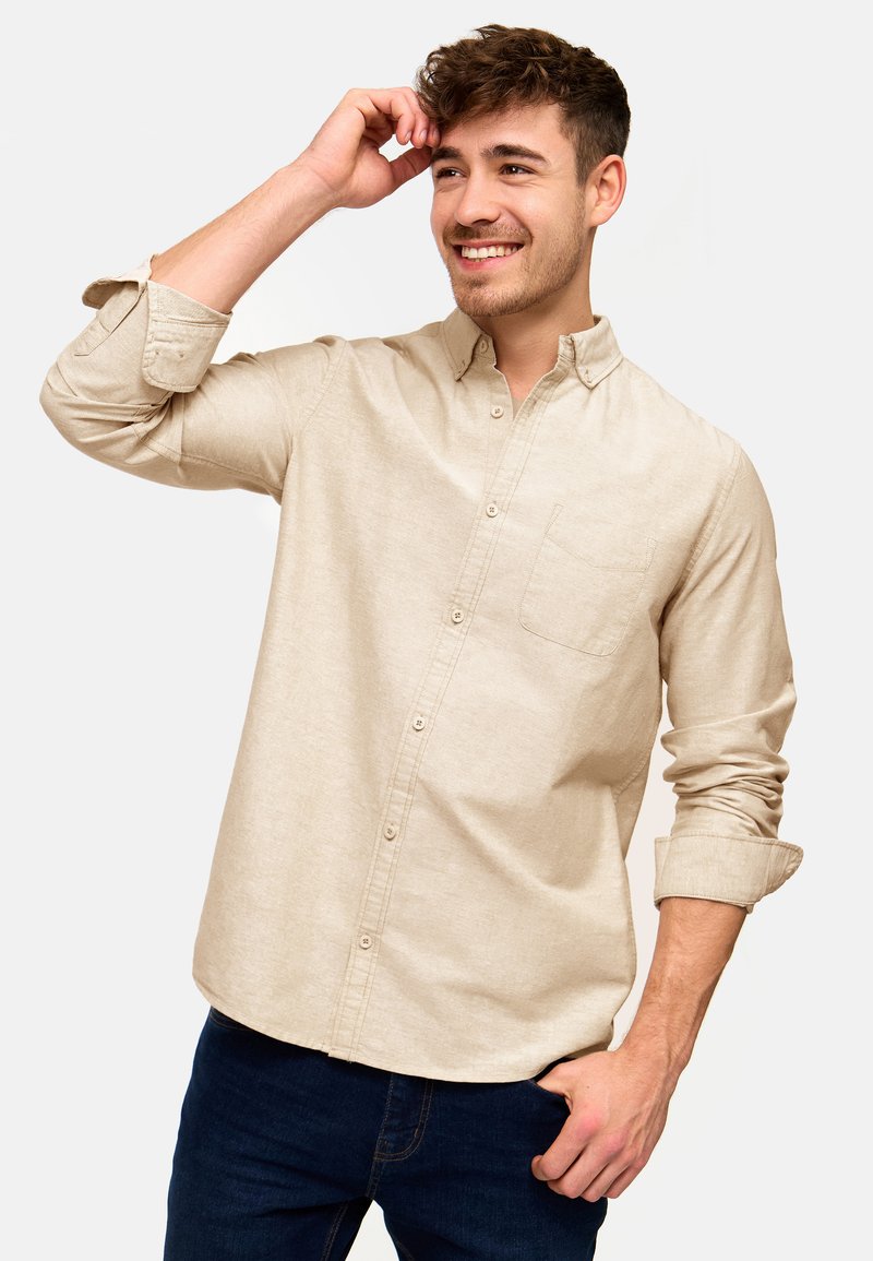Beige Button-up-Hemd mit kleiner Brusttasche, langärmlig und hochgerollt, aus strukturiertem Stoff. Getragen über dunkelblauen Jeans.