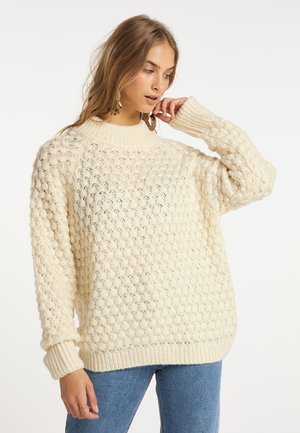 Giovane donna che indossa un maglione lavorato a maglia color crema e jeans blu, che si tocca il viso con una mano, guardando verso il basso.