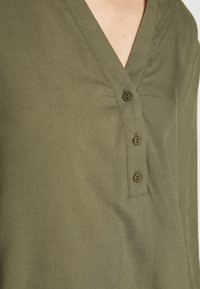 Blouse vert olive avec un col en V, trois boutons au niveau du plastron et un tissu doux et lisse avec un léger éclat.