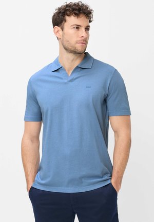 Man with curly hair wearing light blue short-sleeve polo shirt and dark blue pants, hands in pockets, looking to the side.