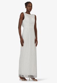 Long white dress with textured, wave-like bead patterns, sleeveless design, and a floor-length silhouette. Paired with strappy white heels.