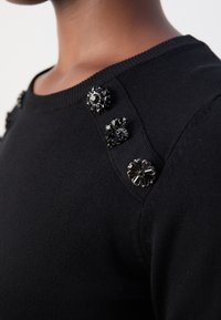 Gros plan d'un haut en tricot noir avec trois boutons décoratifs noirs ornés de bijoux le long de la couture de l'épaule.