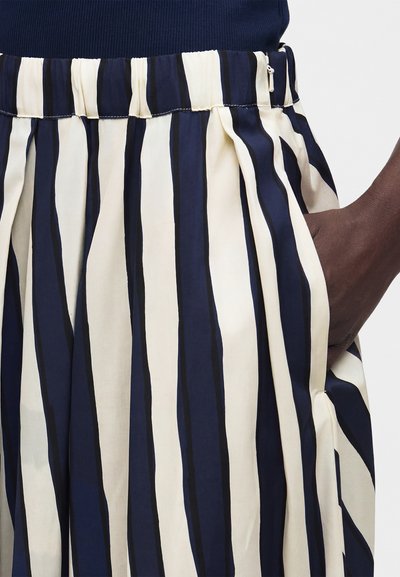 Φούστα μπλε του ναυτικού και λευκές κάθετες ρίγες με ελαστική μέση και πλαϊνό φερμουάρ, φορεμένη με μπλε του ναυτικού τοπ μέσα στη φούστα και χέρι στην τσέπη.