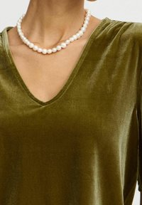 Top in velluto verde oliva con scollatura a V e texture liscia. Una collana di perle bianche riposa sul collo, aggiungendo un contrasto.
