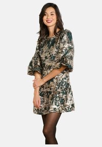 Krátke metalické šaty s kvetinovým vzorom v zelenej a zlatej farbe, s nafúknutými rukávmi a priliehavým pásom, doplnené o priesvitné čierne pančuchy.