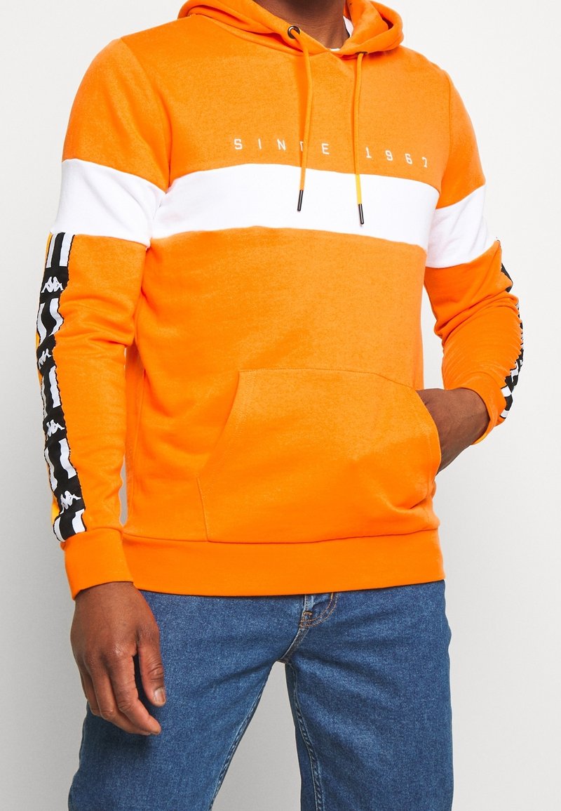 Homme portant un sweat à capuche orange vif avec une bande horizontale blanche et des rayures noires à motifs sur les manches, associé à un jean bleu.