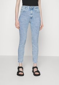 Vaqueros de talle alto en azul claro con un corte slim, que presentan un diseño clásico de cinco bolsillos y un desvanecido sutil. Combinados con sandalias negras.