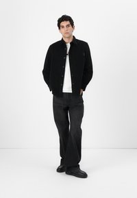 Chaqueta de pana negra con cierre de botón, bolsillo frontal, combinada con una camiseta blanca y jeans de mezclilla negros. El modelo se encuentra de pie con las manos en los bolsillos.
