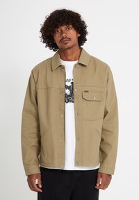 Homme aux cheveux bouclés portant une veste camel à boutons sur un t-shirt blanc avec un motif graphique, debout devant un fond clair uni.