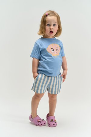 Toddler with short blonde hair, wearing a blue shell-print t-shirt, striped shorts, and pink clogs with charms, standing with hands in pockets.