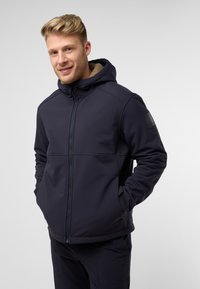 Veste à capuche bleu marine en matériau softshell avec fermeture éclair et doublure en polaire, dotée d'un écusson logo sur la manche.