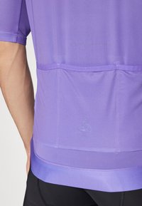 Fialová športová košeľa s textúrovaným sieťovým chrbtom, ergonomickým dizajnom a potlačeným logom na spodnom leme. Bezšvová konštrukcia zvyšuje pohodlie.