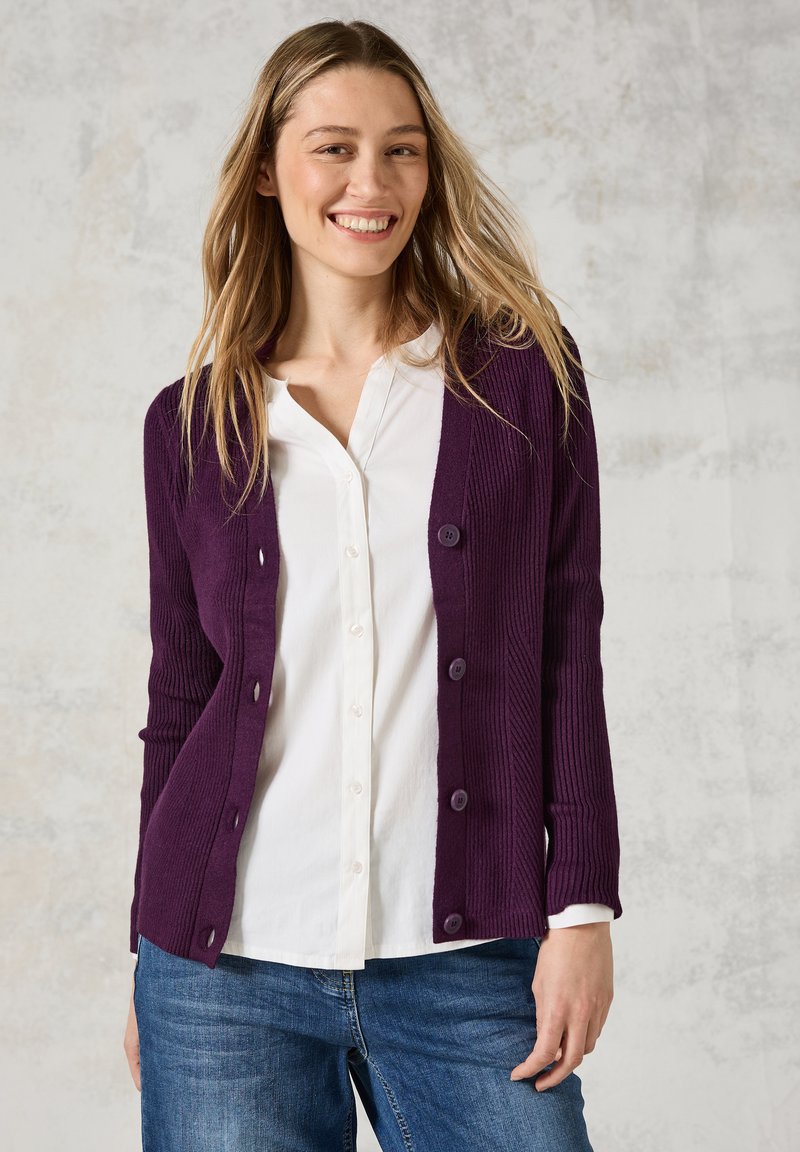 Lila gerippter Cardigan mit Knopfverschluss, strukturiertem Stoff und langen Ärmeln, getragen über einem weißen Hemd mit blauen Jeans.
